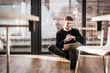 Businessman sitting on floor of his office, using laptop and smartwatch