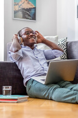Happy young man sitting on the floor in the living room using laptop and headphones