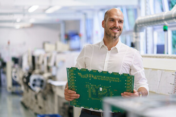 Smiling male technician holding large circuit board while looking away at factory