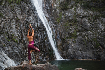 Italy, Lecco, woman doing Tree Yoga Pose on a rock near a waterfall