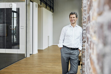 Portrait of smiling businessman leaning against brick wall in modern office