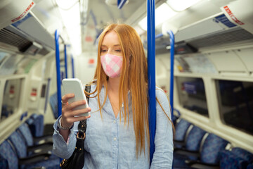 Young woman in mask using smart phone while standing in train