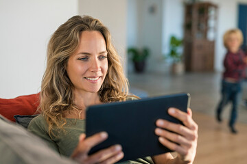 Smiling woman realxing on couch at home using tablet with son in background