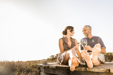 Happy couple sitting on boardwalk at the coast enjoying a glass of white wine