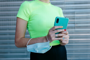 Woman standing in front of metal background, using smartphone, with protective mask on her arm