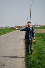 Travel concept. Young man hitchhiker with backpack, stands by rural country road on sunny spring...