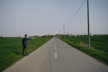 Travel concept. Young man hitchhiker with backpack, stands by rural country road on sunny spring...
