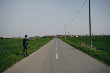 Fototapeta premium Travel concept. Young man hitchhiker with backpack, stands by rural country road on sunny spring day and waiting for car. Front view. Guy went on trip alone, trying to stop passing car. Rear view.