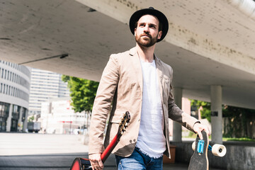 Young man with guitar and skateboard walking in the city