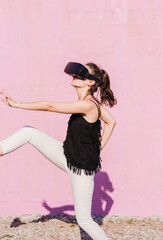 Young woman wearing VR glasses moving in front of pink wall