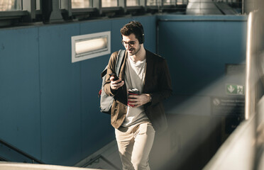 Smiling young man with headphones, cell phone and takeaway coffee walking at the station