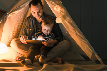 Father and son sharing a tablet in a dark tent at home