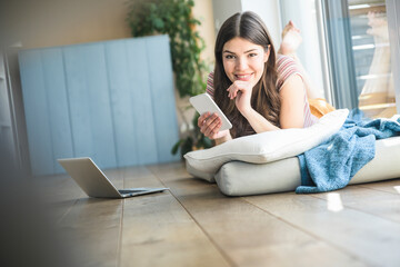 Portrait of smiling young woman with cell phone lying at the window at home