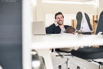 Young businessman sitting in office, with feet on desk, talking on the phone