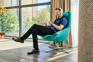 Young man sitting in armchair, listening music, using digital tablet