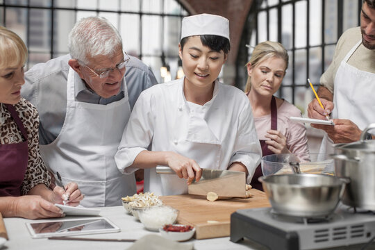 Participants in cooking class watching female chef chopping ingredients