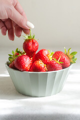 A female hand takes a ripe red strawberry from a green bowl on a gray table. Concept of summer food. Vertical orientation. Selective focus.