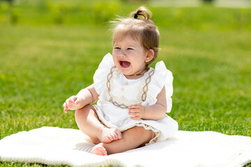 A baby in a white dress sitting on the grass and crying. crying baby toddler girl on grass outdoors. Child feelings and emotions concept. Sad expression. Childhood. copy space