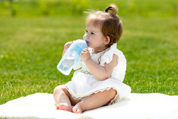 A cute toddler girl in a white summer dress sitting on the grass in the summer, drinking water from a bottle. Healthy toddler one year sitting on the grass drinking water from a bottle