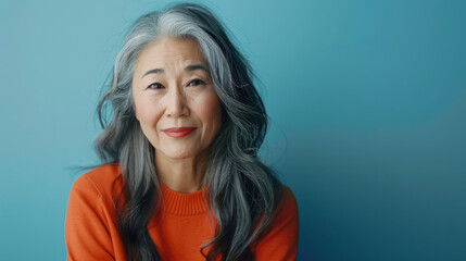 Senior Asian woman with long grey hair smiling, wearing an orange sweater on a blue background