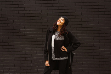 Young woman leaning back, smiling and looking up, standing against a black brick wall on the street.