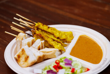 Grill pork satay with peanut sauce, grilled bread and cucumber salad in round shape paper plate on wooden table. Food popular Thai food snack called Moo Satay, the pork skewers with peanut sauce.
