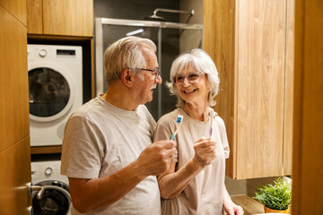 Happy senior couple standing in bathroom with toothbrushes in hands and smiling at each other.