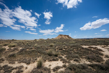 A wide view of the desert landscape in Las Bardenas Reales, Spain, under a clear blue sky with scattered clouds. Sparse vegetation and rugged terrain are prominent.