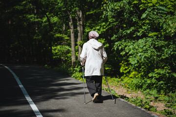 An elderly woman is leisurely walking along a road, aided by walking sticks, in a natural setting...