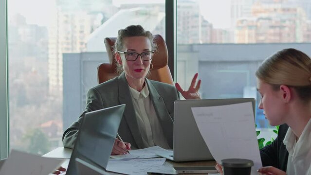 A confident middle-aged blonde girl with glasses and a business gray suit speaks to her businesswoman colleagues while sitting at a table with laptops and important papers in the office