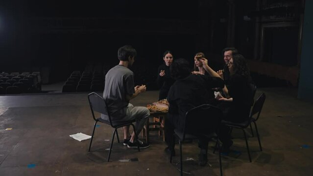 A group of happy actors in black uniforms sit around an impromptu pizza and chat during a break and preparation for a performance on stage in a theater