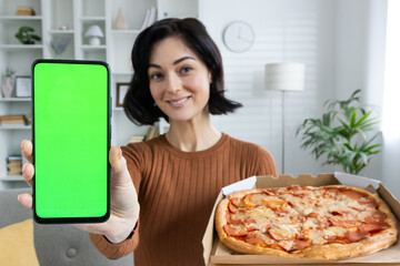 Close-up portrait of a young smiling girl advertising a food delivery app. Standing at home, holding a pizza box and showing the green screen of a mobile phone to the camera