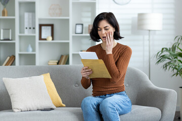 Upset and worried young woman reading a received letter at home, sitting on the sofa and holding...
