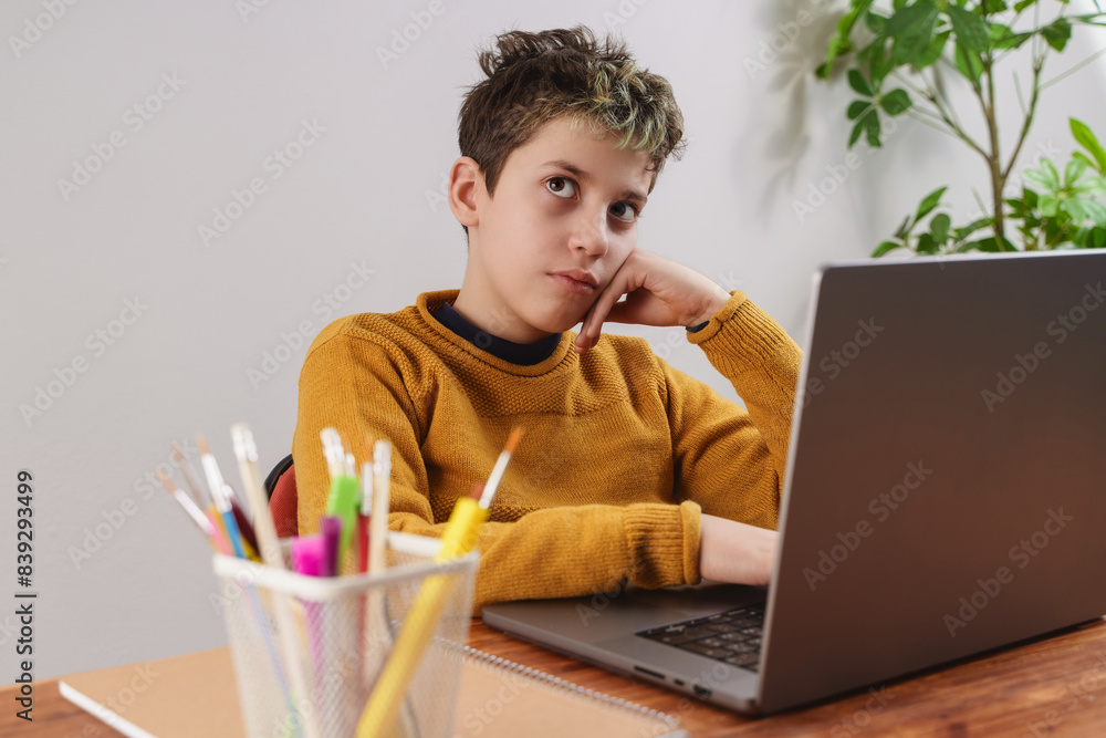 Wall mural distracted boy studying on laptop sitting at desk - contemplative student during online learning