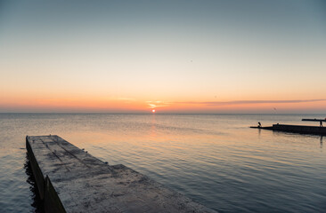 Sea coast with few piers and silhouettes of fishermen at sunrise. Calm water, seagulls are resting on the concrete pier, sunrise over the sea in the background. 