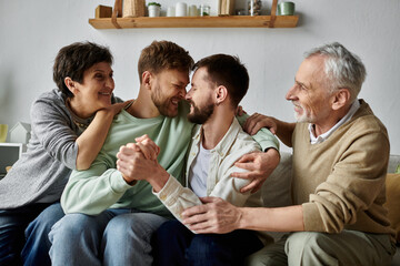 A gay couple sits on a couch with parents, all smiling and embracing each other.