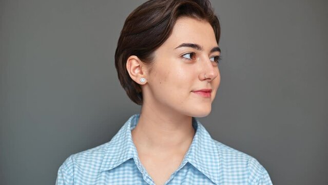 Portrait of smiling cute young brunette woman with short haircut in blue shirt turning around and looking at camera standing on grey background