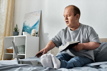 A man with inclusivity sits in bed, working on a laptop and holding a book.