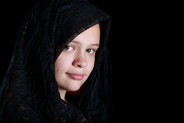 A pale skinned teen girl wears a black shawl over her head looking at the camera.