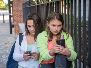 Two girls are sitting on a bench and looking at their cell phones