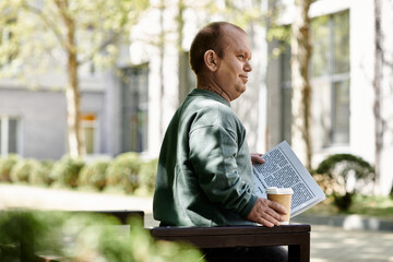 A man with inclusivity sits on a bench in a park, reading a newspaper and holding a cup of coffee.