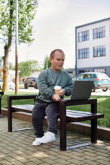 Fototapeta premium A man with inclusivity sits on a bench in a park, enjoying a cup of coffee and working on his laptop.