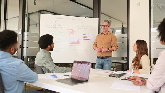 A charismatic mature male presenter leads a dynamic brainstorming session in a contemporary office setting, diverse team listens intently. A vibrant exchange of ideas and a culture of collaboration