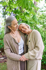 Fototapeta premium Two middle-aged women, wearing cardigans, embrace each other lovingly under the shade of green trees.