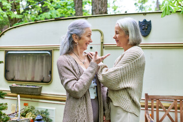 Two middle-aged women, a lesbian couple, chat and laugh while camping in a forest setting.