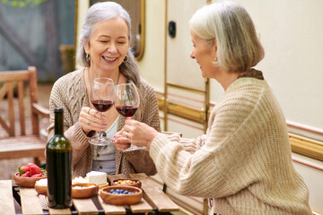 Two middle-aged women raise a toast with wine glasses while camping in a lush green forest.