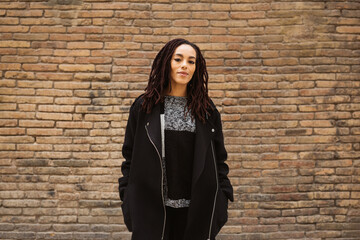 Portrait of a young beautiful woman with dreadlocks standing against a brick wall on the street.