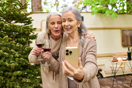 Two middle-aged women enjoying wine and taking a selfie while camping in a forest.