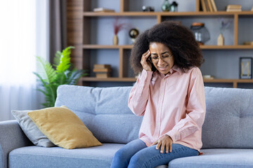 Obraz premium Young woman in glasses sitting on a cozy sofa, holding her head in pain, indicating a headache or stress in a contemporary living room setting.