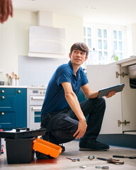 Male Plumber With Digital Tablet Fixing Waste Disposal Unit In Domestic Kitchen Sink